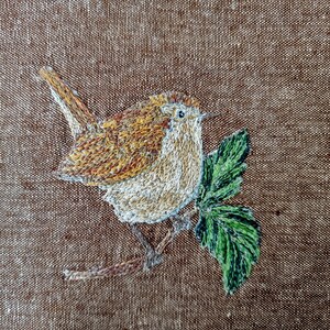 May include: Embroidered artwork of a brown wren perched on a green leaf branch. The bird is stitched with brown, white, and orange thread. The leaf is stitched with green thread.