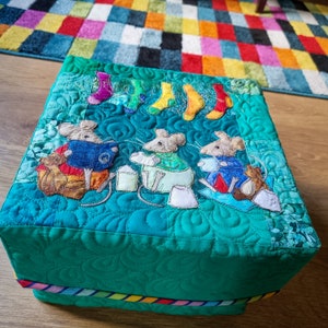 May include: A square, quilted box in shades of teal and green, adorned with appliqued mice and colorful socks. The box features a whimsical design with embroidered details and a rainbow-striped trim at the base. The top is decorated with three mice and socks.