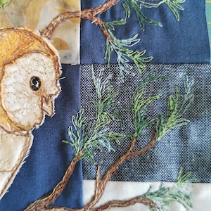 May include: A close-up of a hand-stitched fabric art piece depicting a barn owl with brown and white feathers, perched on a branch of a pine tree with green needles. The background is a patchwork of blue and white fabrics.