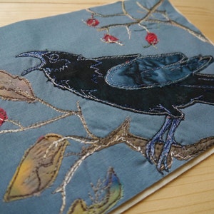 May include: A blue fabric bird with black and white accents is stitched onto a blue fabric background. The bird is perched on a branch with brown leaves and red berries.