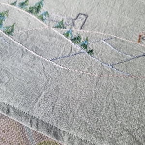 Puede incluir: Primer plano de una tela gris con detalles bordados de una cordillera con árboles y casas. La tela tiene una superficie texturizada y un borde cosido.