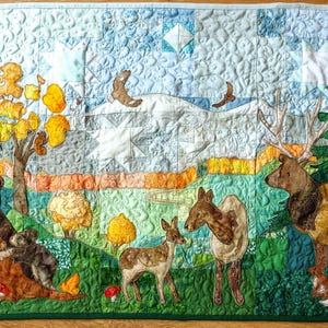 May include: A quilted wall hanging depicting a forest scene with a bear family, deer, and a fox. The artwork features trees, a mountain range, and birds in flight. The color palette includes greens, browns, yellows, and blues.