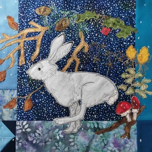 May include: A white rabbit with long ears is embroidered on a blue fabric with white polka dots. The rabbit is surrounded by brown branches, green leaves, and red berries. The fabric is part of a larger quilt with blue and white squares.