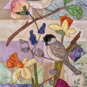 Könnte beinhalten: Ein gequiltetes Wandbehang mit zwei Vögeln, die auf Ästen mit Blumen und Blättern sitzen. Die Vögel sind grau und weiß mit schwarzen Augen. Die Blumen sind gelb, orange, rosa und lila. Die Blätter sind grün.