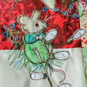 May include: A white mouse with a green and blue waistcoat is embroidered on a white fabric background. The mouse is holding a green flower with white petals. The background is a red fabric with a floral pattern.