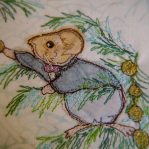 Peut inclure: Image brodée d'une petite souris portant un costume bleu et tenant un ornement rose. La souris est entourée de feuillage brodé vert et bleu.