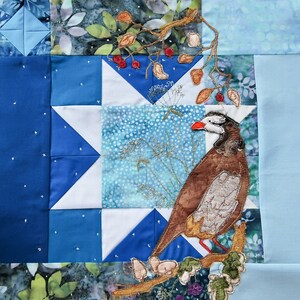 May include: A blue and white patchwork quilt with a brown and white bird embroidered on it. The bird is perched on a branch with red berries. The quilt is made of different fabrics and has a rustic look.