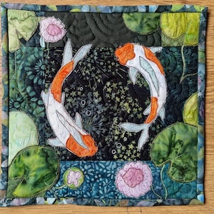 Könnte beinhalten: Ein quadratisches Quilt mit zwei Koi-Karpfen in einer Teichszene. Die Fische sind orange und weiß, vor einem dunklen Hintergrund mit Seerosenblättern und rosa Blüten. Der Rand besteht aus einer Mischung aus grünen und blauen Stoffen.