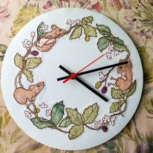 May include: A white circular clock with a black clock face and red clock hands. The clock face is surrounded by a wreath of embroidered green leaves, brown mice, and white flowers.
