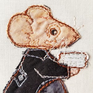 May include: A brown and white mouse wearing a black jacket and holding a white cup. The mouse is embroidered on a white fabric background.