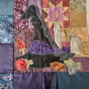 May include: A quilted fabric wall hanging featuring a black witch's hat, purple and gold candles, and red and white mushrooms. The background is a patchwork of different fabrics in shades of purple, brown, and gold.