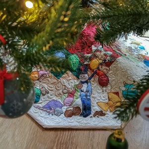 May include: A white quilted Christmas tree skirt with a colourful embroidered design featuring a nutcracker, a bow, and other festive elements. The skirt is decorated with a red ribbon and a silver bauble.