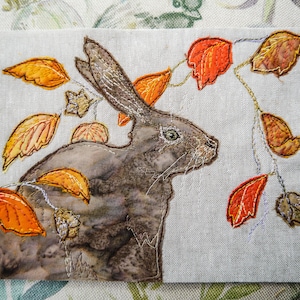 May include: A brown rabbit with a white patch on its chest is surrounded by orange and brown leaves. The rabbit is embroidered on a white linen fabric.