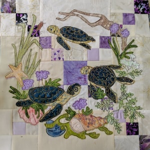 May include: A quilted fabric wall hanging with an embroidered design of three sea turtles, a starfish, and various sea plants. The turtles are blue and green with gold accents. The background is a patchwork of purple and white squares.