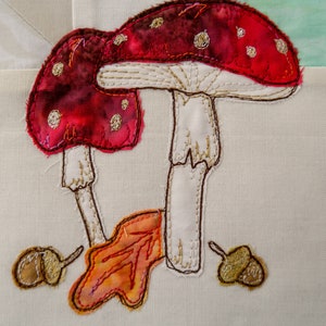 May include: Embroidered fabric art featuring two red and white mushrooms with brown spots, a brown leaf, and two acorns. The mushrooms have white stems and are stitched with brown thread.