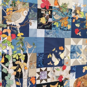May include: A patchwork quilt with appliqued images of woodland creatures and foliage. An owl perches on a branch, while birds and a deer are also depicted. The quilt has a blue, beige, and white colour scheme.