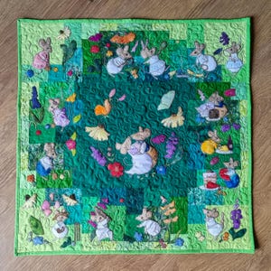 May include: A square, quilted wall hanging featuring a whimsical design of mice in a garden setting. The quilt has a green background with various shades of green squares. The mice are dressed in various outfits and surrounded by flowers and butterflies.