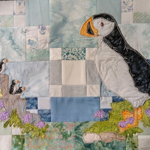 May include: A quilted wall hanging featuring a white, black, and orange embroidered puffin with a green and yellow floral background.