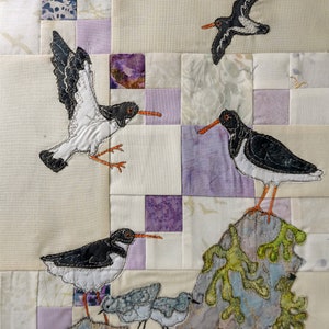 May include: A quilt block with a patchwork design featuring four black and white birds embroidered on a cream background. The birds are perched on a rock with green seaweed and a purple jellyfish.