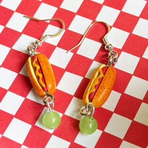 May include: A pair of silver dangle earrings with a hot dog charm and a green bead. The hot dog charm is red, yellow, and brown.