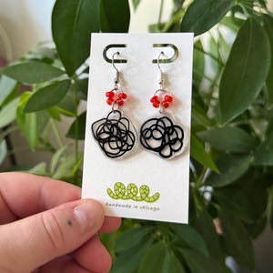 Puede incluir: Un par de pendientes colgantes con diseños de garabatos negros sobre un fondo transparente. Los pendientes están adornados con cuentas rojas y ganchos plateados. La tarjeta dice "hecho a mano en Chicago".