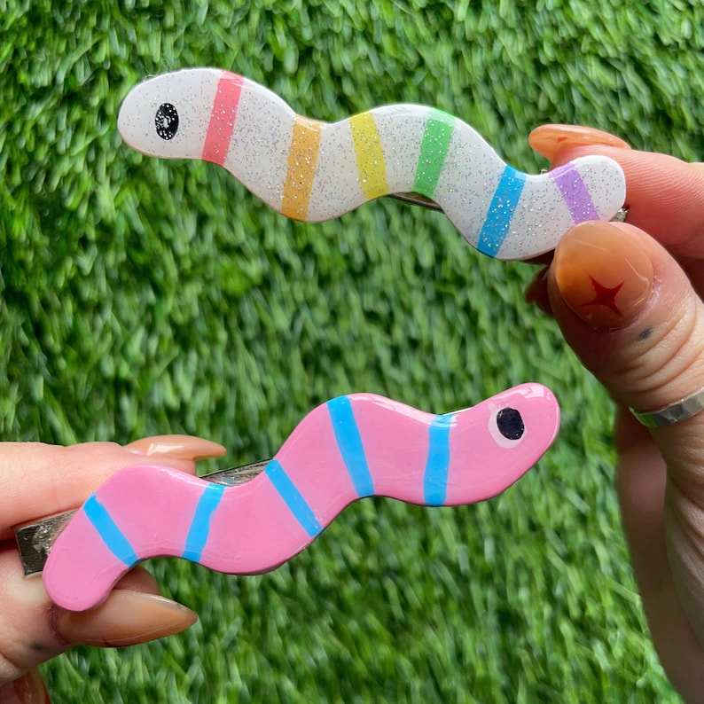 May include: Two colourful hair clips shaped like worms. One is pink with blue stripes and the other is white with rainbow stripes.