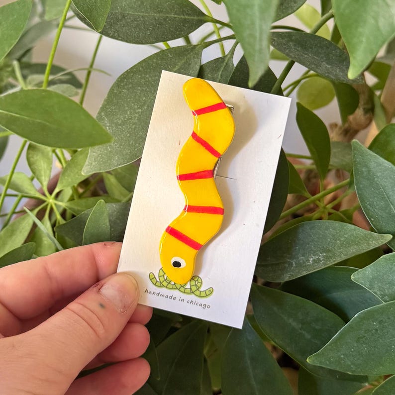 May include: A yellow and red ceramic worm pin with a green worm in its mouth. The pin is on a white card with the text "handmade in chicago".