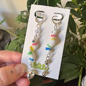 以下が含まれることがあります： 銀色のぶら下がりイヤリングで、白地にカラフルなストライプの入ったミミズと、ディスコボールのチャームが付いています。イヤリングは、白いカードに「Handmade in the」という文字と、緑色のミミズのデザインが描かれています。