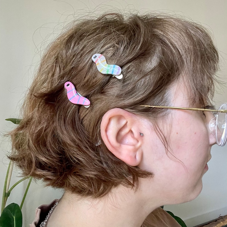May include: A person with short brown hair wearing glasses has three colourful hair clips in their hair. The clips are shaped like a boomerang and have a striped pattern.