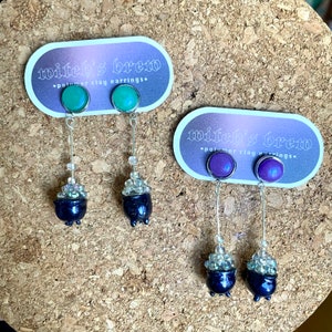 Könnte beinhalten: Zwei Paare Hexenbrau-Ohrringe aus Polymer Clay. Das erste Paar hat grüne, irisierende Cabochons mit silberfarbenen Ohrhaken und schwarzen Kesselanhängern mit weißen Perlen. Das zweite Paar hat violette, irisierende Cabochons mit silberfarbenen Ohrhaken und schwarzen Kesselanhängern mit weißen Perlen.