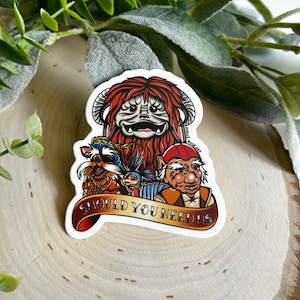 May include: A sticker featuring a group of characters from the movie Labyrinth. The characters include a furry red creature with a large head, a small blue creature with a pointed hat, a dog-like creature with a brown and white coat, and a human-like creature with a red hat and a long white beard. The sticker has the text "Should You Need Us" on a banner.