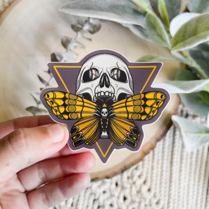 May include: A sticker featuring a black and white skull with a yellow and black butterfly emerging from its mouth. The skull and butterfly are set against a purple background with a yellow triangle border.