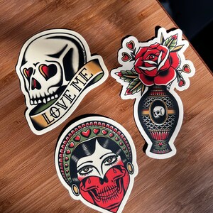 May include: Three vintage-style tattoo flash designs. The first is a skull with hearts for eyes and a banner that reads "Love Me". The second is a woman with a skull for a face and a crown of hearts. The third is a rose in a vase with a skull on the side.