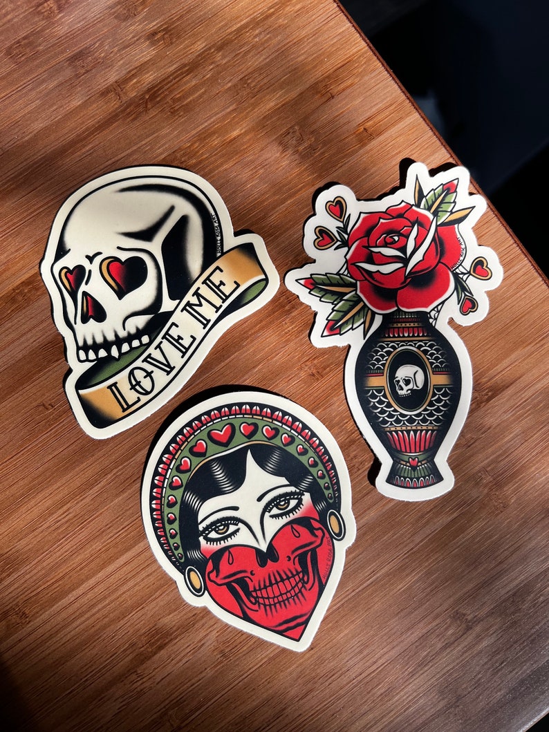 May include: Three vintage-style tattoo flash designs. The first is a skull with hearts for eyes and a banner that reads "Love Me". The second is a woman with a skull for a face and a crown of hearts. The third is a rose in a vase with a skull on the side.
