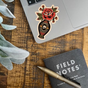 May include: A vintage style sticker of a red rose in a vase with a skull design. The vase is decorated with a geometric pattern and the rose has a few hearts around it.