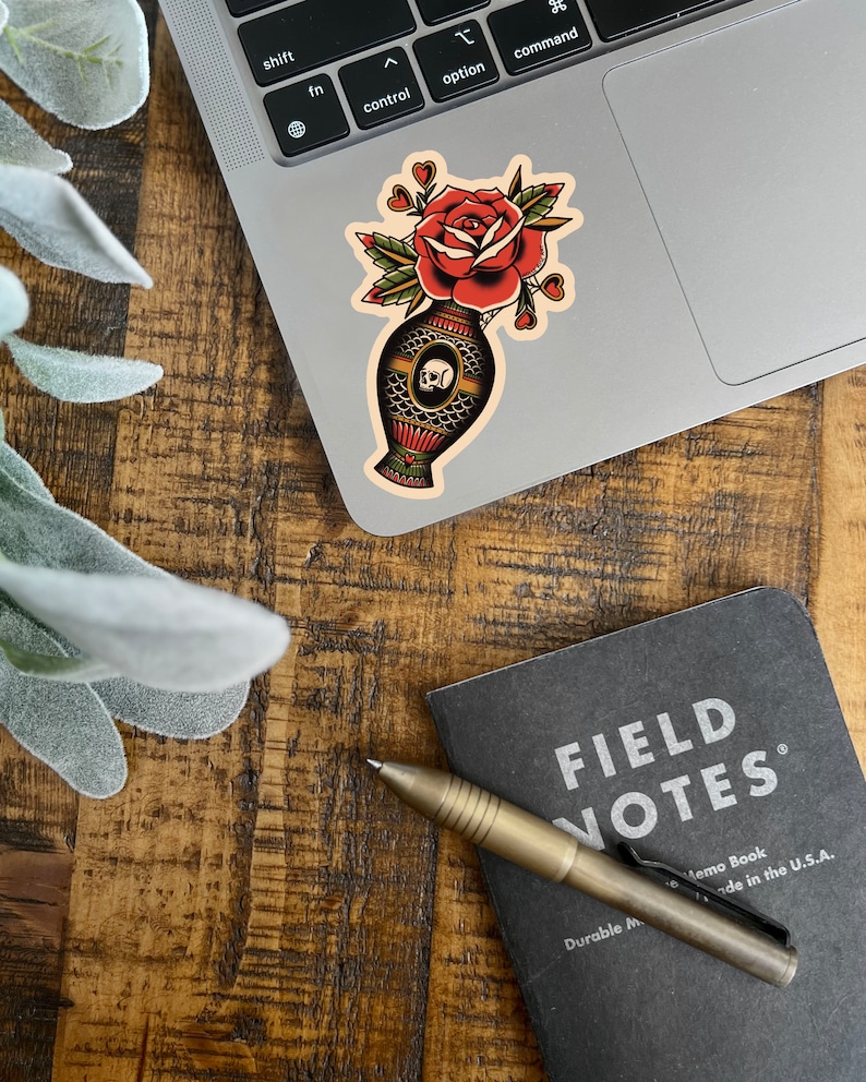 May include: A vintage style sticker of a red rose in a vase with a skull design. The vase is decorated with a geometric pattern and the rose has a few hearts around it.