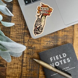 May include: A sticker with a skeleton hand holding another hand, with the text "Partner in Crime" on a banner. The sticker is on a silver laptop.