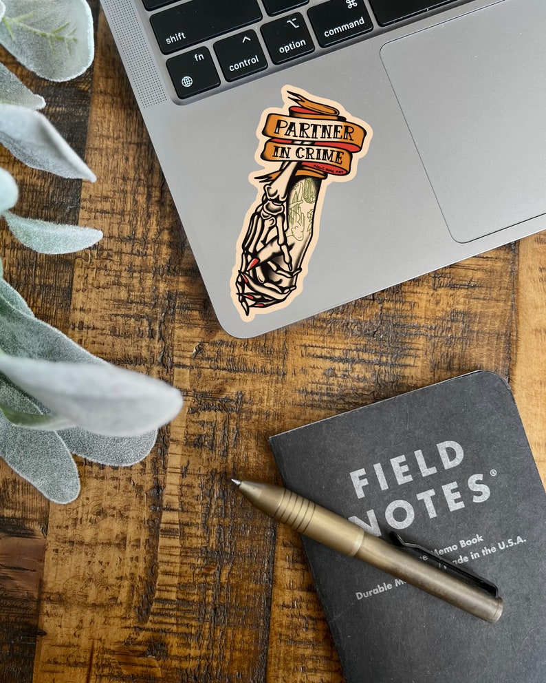 May include: A sticker with a skeleton hand holding another hand, with the text "Partner in Crime" on a banner. The sticker is on a silver laptop.