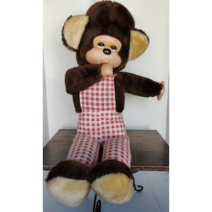 May include: A vintage plush monkey doll with a brown fur body, large ears, and a red and white checkered overall. The monkey has a painted face with a neutral expression and is seated with its arms outstretched. The doll is a nostalgic toy.