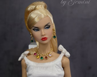 May include: A blonde doll wearing a white top, red belt, and a black and white checked skirt. She is wearing a gold necklace with colourful gemstones and matching earrings. The doll has a gold bracelet with colourful gemstones on her wrist.