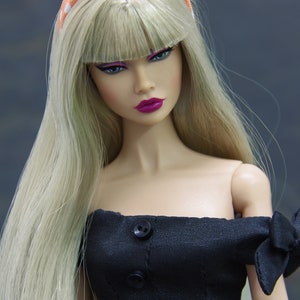 May include: A doll with long blonde hair wearing a black off-the-shoulder dress with buttons down the front and a polka dot headband. The doll has a pink lipstick and a neutral makeup look.