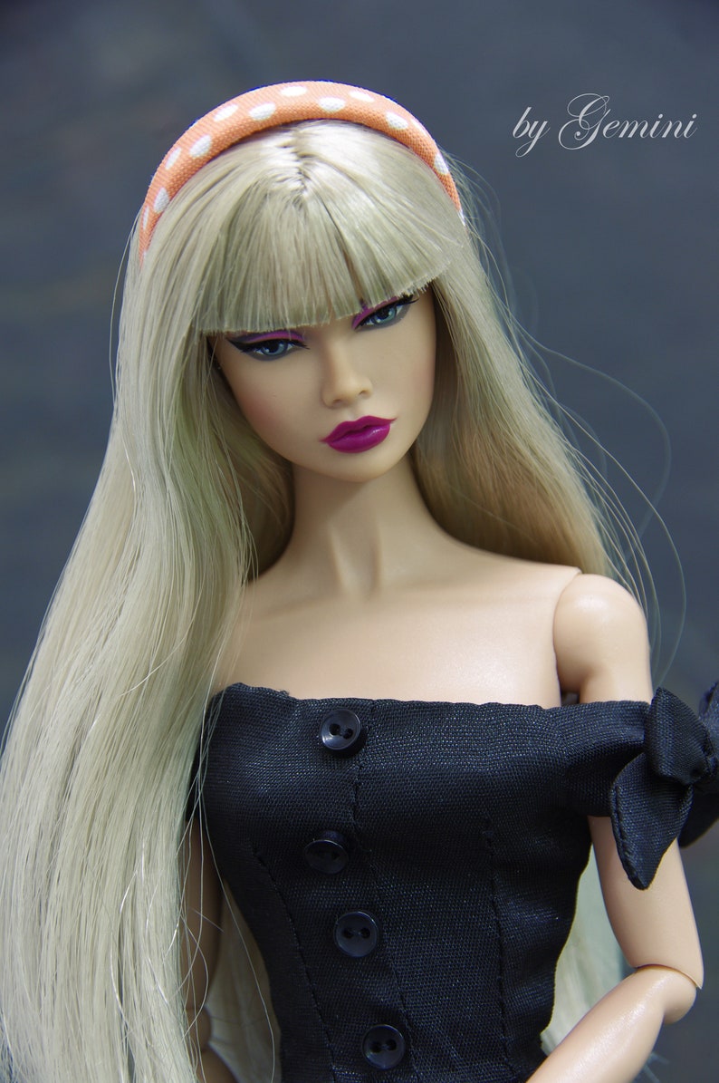 May include: A doll with long blonde hair wearing a black off-the-shoulder dress with buttons down the front and a polka dot headband. The doll has a pink lipstick and a neutral makeup look.