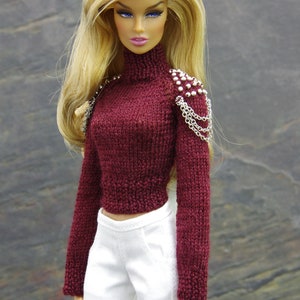 May include: A doll wearing a maroon knitted sweater with silver chain and bead embellishments on the shoulder, white pants, and pink nail polish. The doll has long blonde hair and blue eyes. The text "by Gemini" is in the upper right corner of the image.