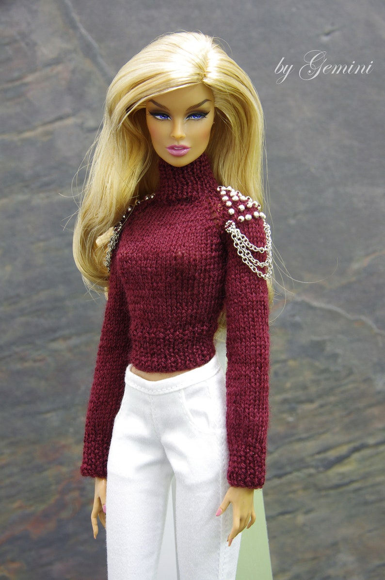 May include: A doll wearing a maroon knitted sweater with silver chain and bead embellishments on the shoulder, white pants, and pink nail polish. The doll has long blonde hair and blue eyes. The text "by Gemini" is in the upper right corner of the image.