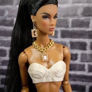 May include: A fashion doll with long black hair in a high ponytail, wearing a cream-colored bustier, and dark denim pants. The doll is adorned with gold jewelry, including a chain necklace and earrings with rectangular pendants. The background is a gray brick wall.