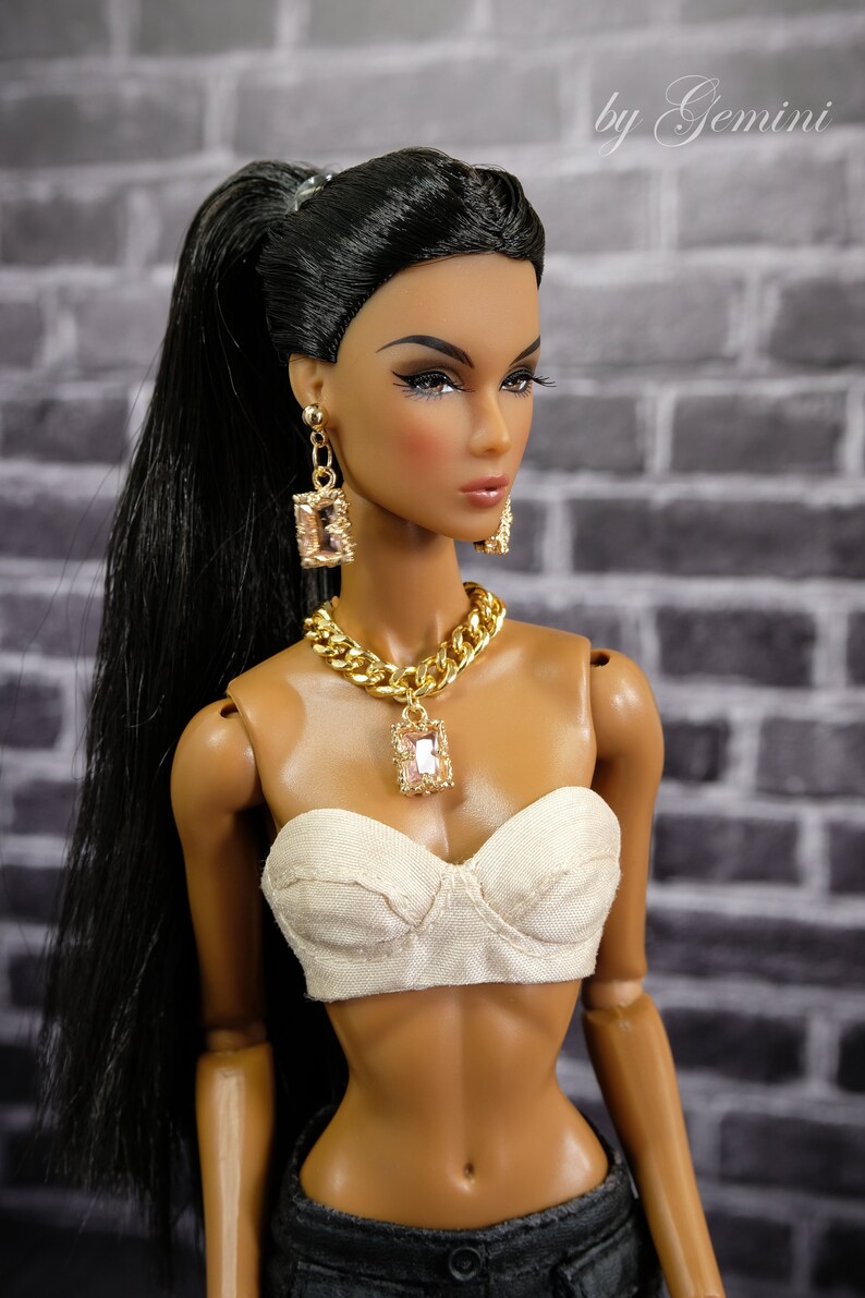 May include: A fashion doll with long black hair in a high ponytail, wearing a cream-colored bustier, and dark denim pants. The doll is adorned with gold jewelry, including a chain necklace and earrings with rectangular pendants. The background is a gray brick wall.