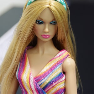 May include: A doll with long blonde hair wearing a colorful striped dress and a green and white polka dot headband.