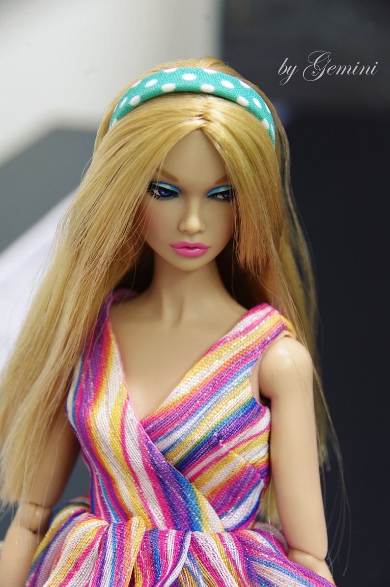 May include: A doll with long blonde hair wearing a colorful striped dress and a green and white polka dot headband.