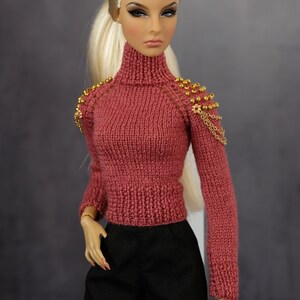 May include: A doll wearing a pink knitted sweater with gold bead and chain embellishments on the shoulders. The doll has blonde hair and is wearing black pants.