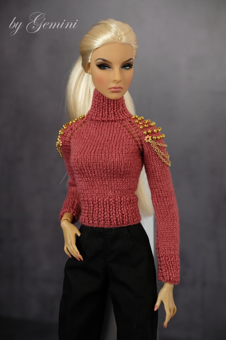 May include: A doll wearing a pink knitted sweater with gold bead and chain embellishments on the shoulders. The doll has blonde hair and is wearing black pants.